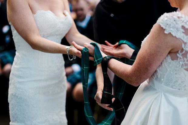 two women dressed in white wedding dresses being handfasted with tartan ribbons.